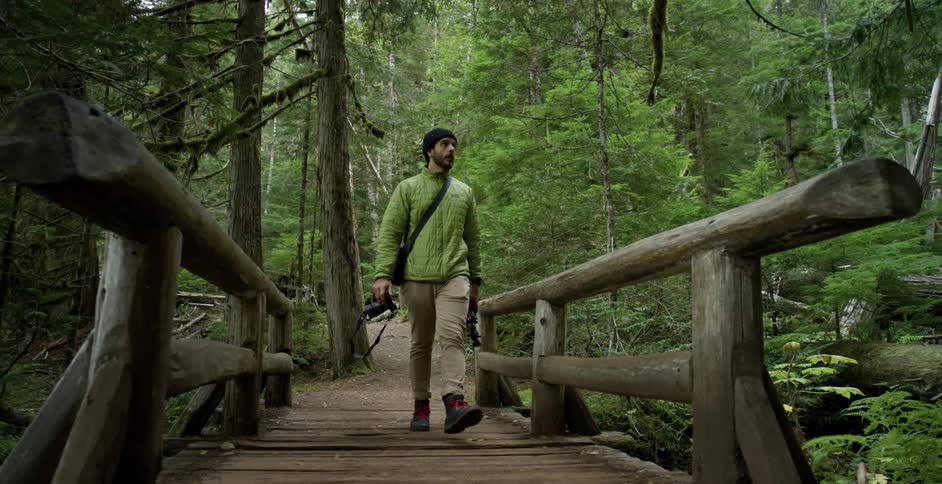 A white man with a camera and tripod crosses a wooden bridge in the forest