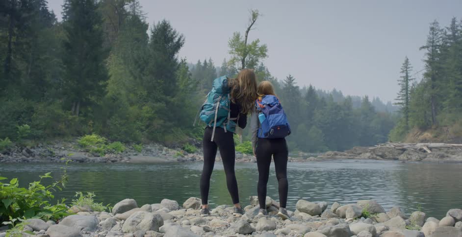 Two girls with backpacks stand on the shore of a lake near pine tree forest