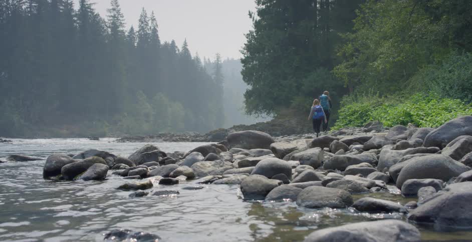 Two white women with backpacks walk along a rocky river shore
