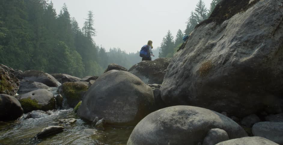 A white woman jumps across big rocks in a stream bed