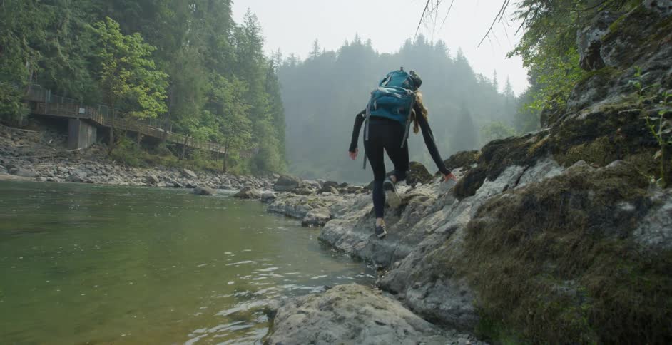 A white woman carefully walks on the rocky shore of a river