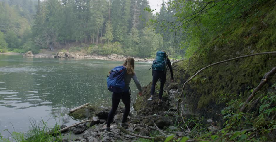 Two women hike over a rocky river shoreline