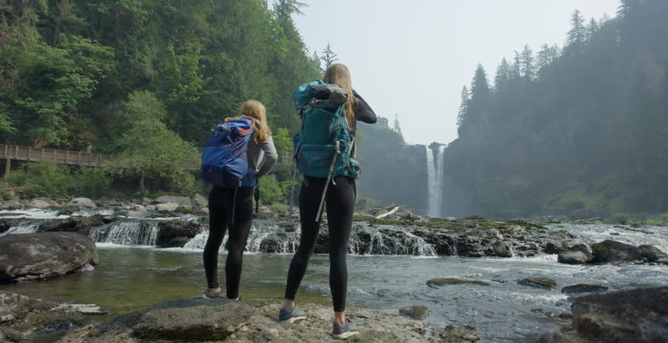 Hikers stand on river shore viewing tall waterfall near bridge and mountains