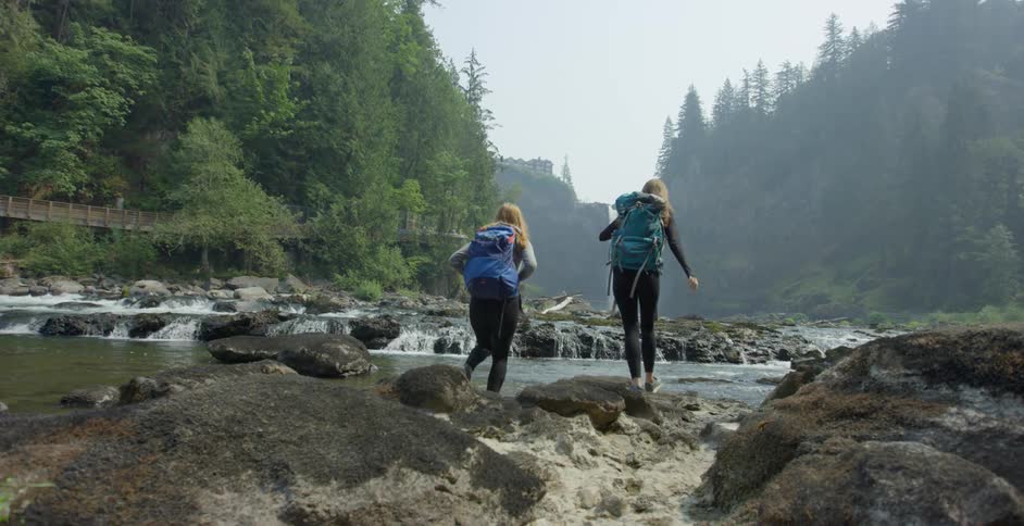 Two women hiking near the river with trees and the sky and a waterfall in the background