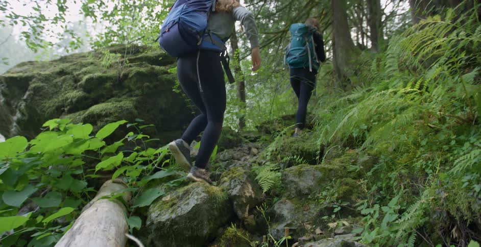 A white woman carefully crosses a log and walks up a hill after her friend