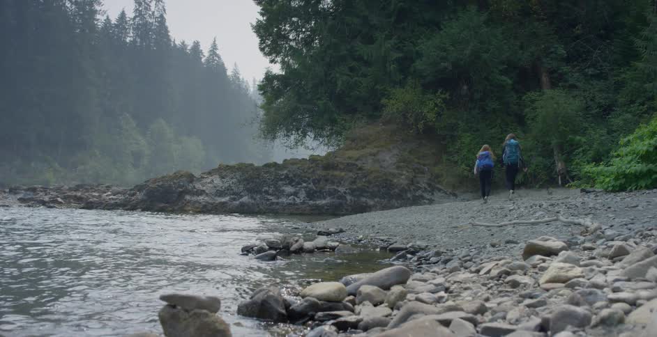 Two white woman hike along a rocky river shoreline