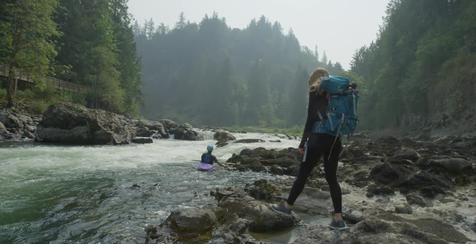 A white woman with a backpack walks over rocks on a river shoreline towards a person sitting in a kayak