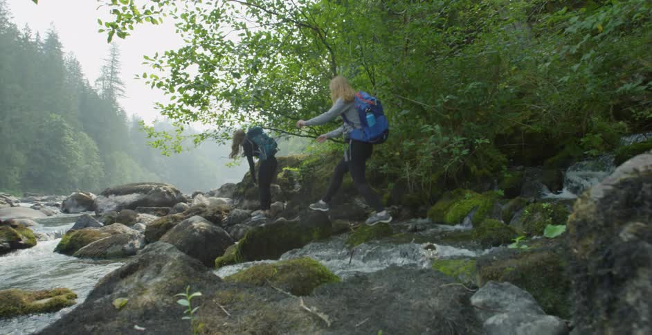 Two girls wearing backpack walk over the rocks of a river in the forest