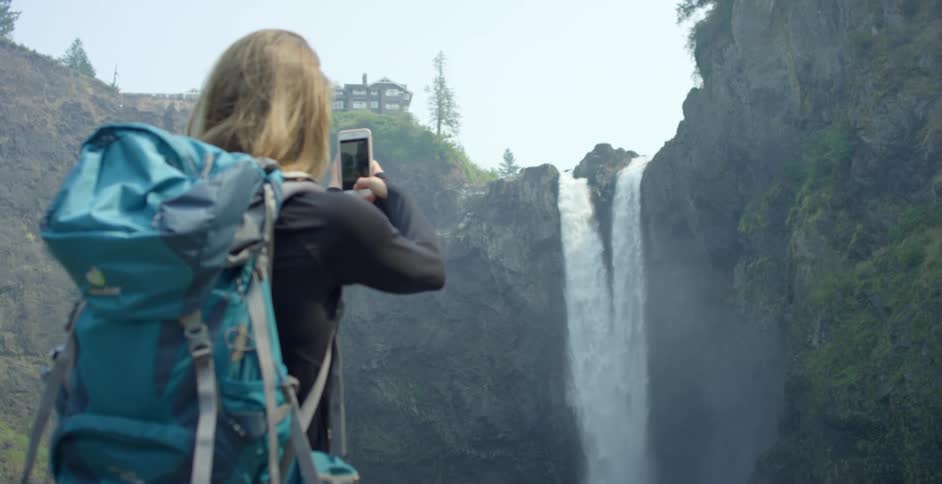 Woman wearing backpack photographs waterfall using mobile phone