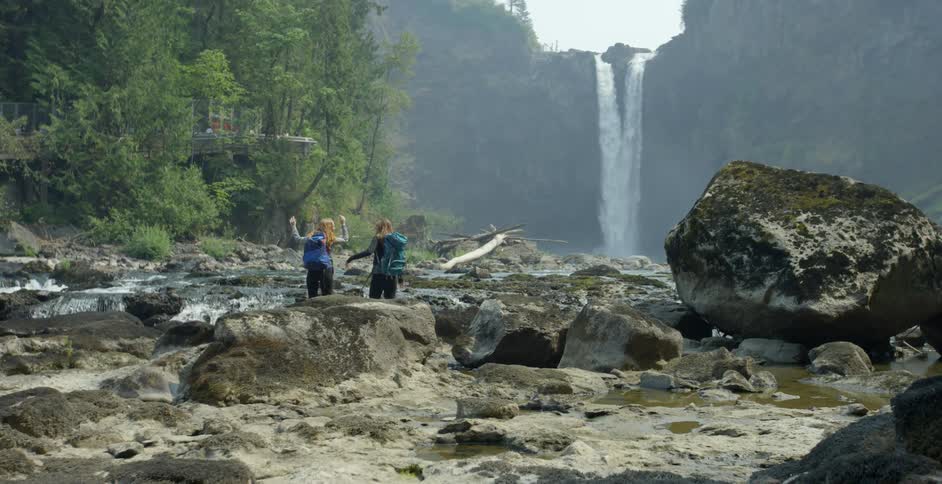 Two girls raise their hands above their heads and hug as they stand in front of a waterfall