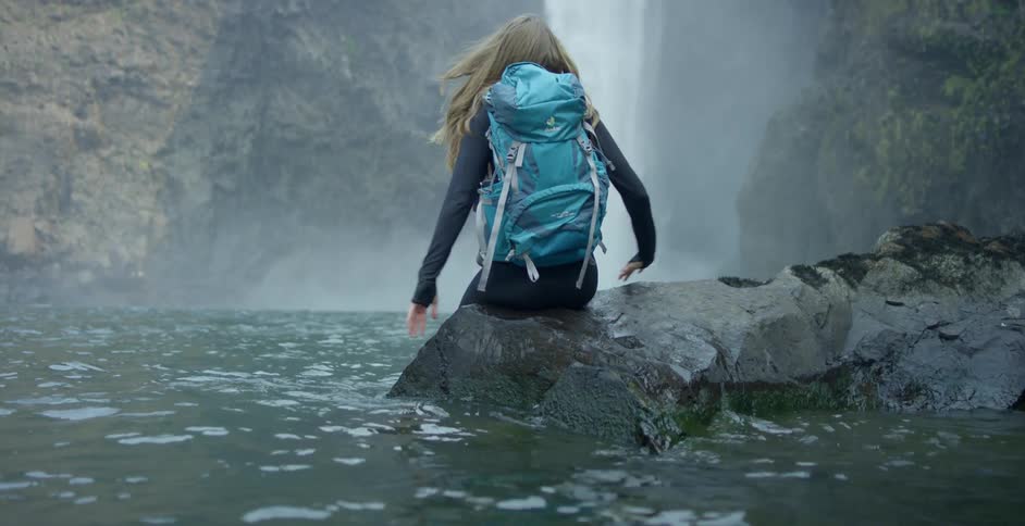 Young girl with backpack sits on a rock in a river in front of a waterfall
