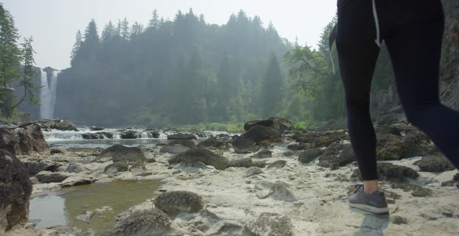 Young girl with backpack walks on the rocky shores of a river near a waterfall
