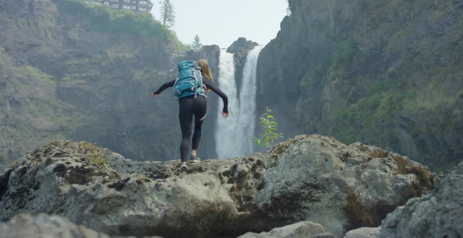 A woman hiking climbs up a rock in front of a water fall