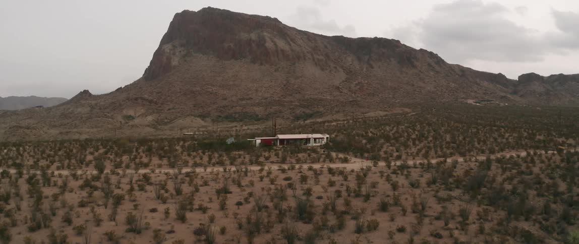 Dirt road passes by house surrounded by scrub land with mountain in background