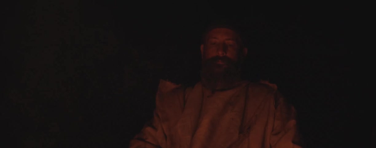 A white man sitting in front of a fire thinking