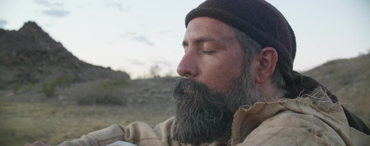 A man wearing a head wrap and tattered robe sits in the desert with eyes closed meditating