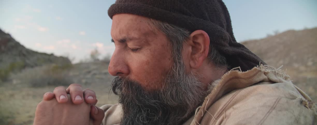 Man with a beard and a brown beanie clasps hands and prays with eyes closed