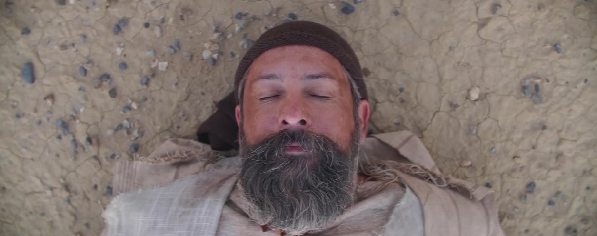 Man with beard wearing head covering lies down on ground sleeping