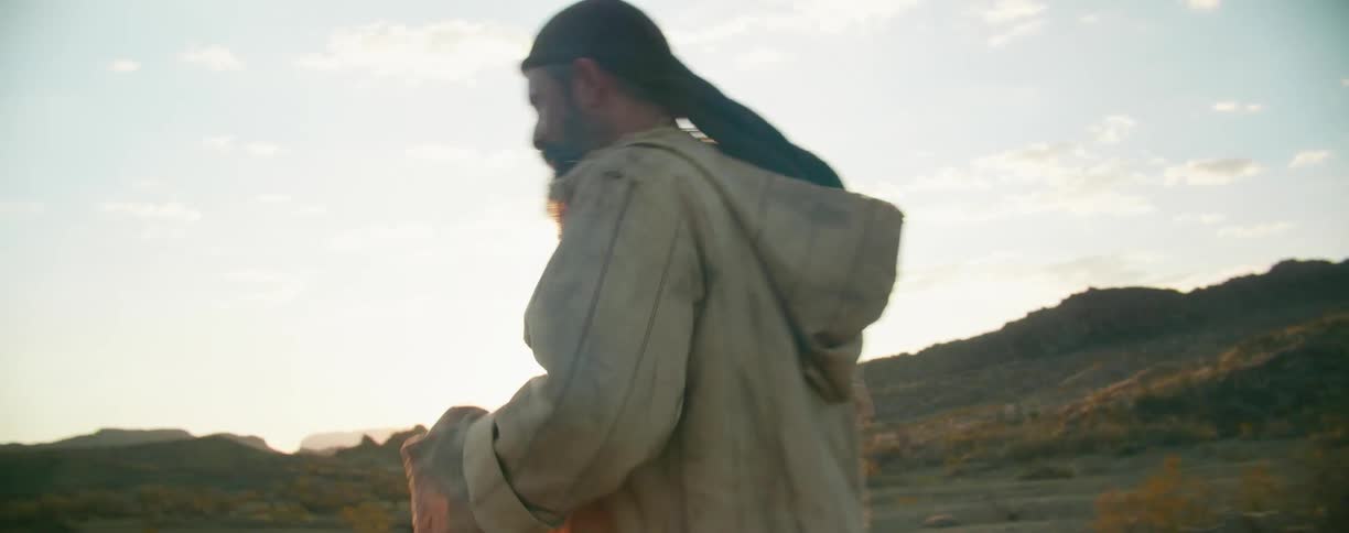 A man wearing a head wrap walks and stumbles in the desert with the sun glaring