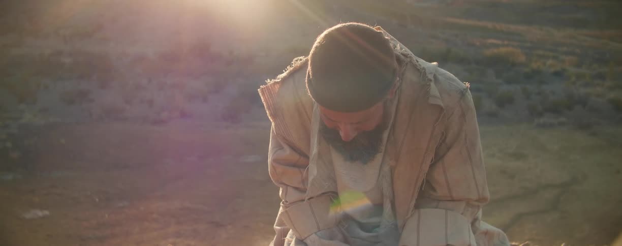 Man nods his head as he kneels on the ground in a field under the bright sun