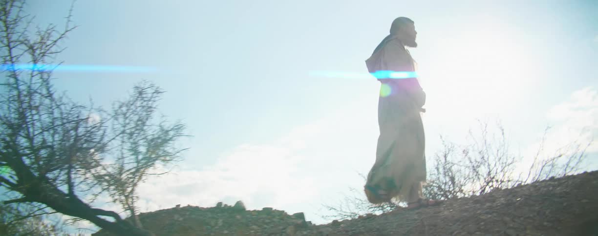 The sun shines and glares down on a man walking along a ridge wearing a robe and sandals