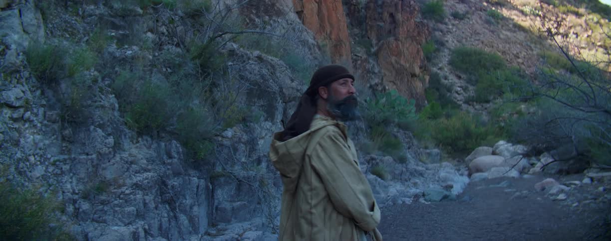 Man wearing a robe walks on a dirt path near a mountain in the desert