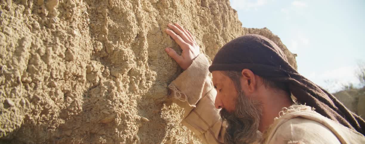 A man stands outdoors with his eyes closed and a hand placed against a large stone