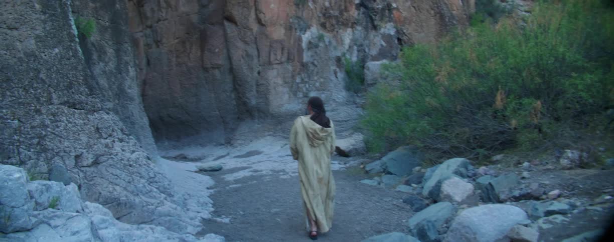 A man in a cloak walks at the base of a canyon