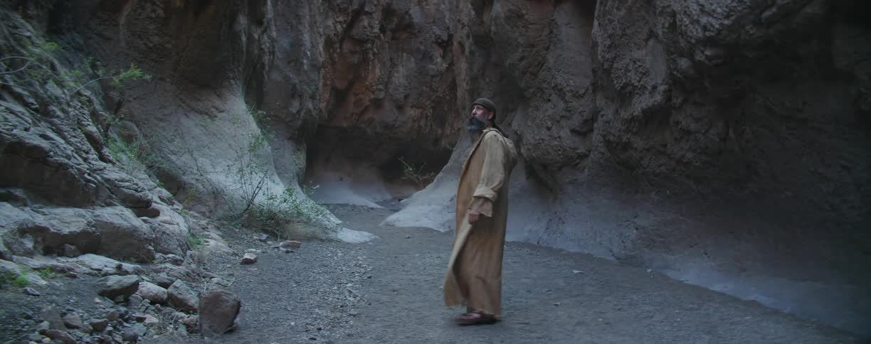 A man in a long cloak walks at the base of a canyon and looks around