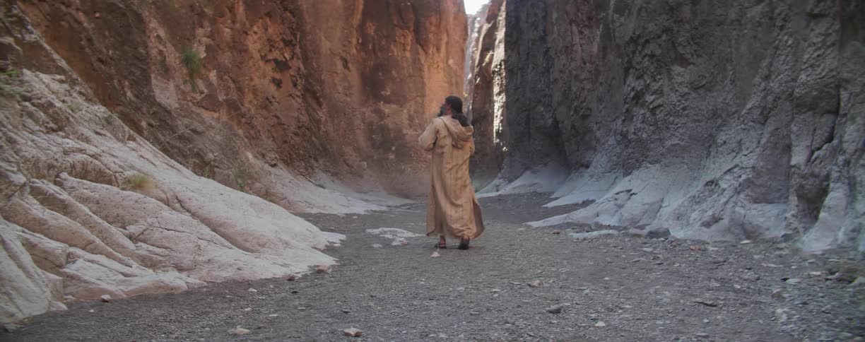 A man with a bandana and a coat walking in the bottom of a canyon