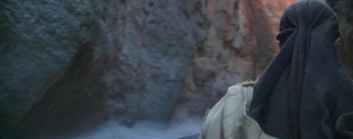 A man dressed in a tunic is walking through a narrow canyon during the day