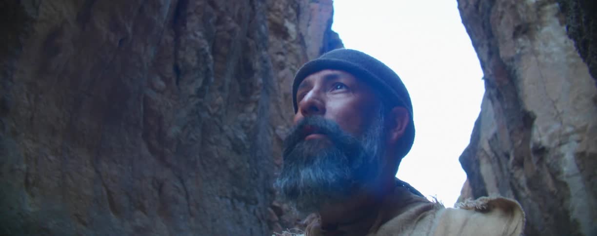 A man with a full beard and mustache wearing a head wrap looks around at the rock walls and then walks off