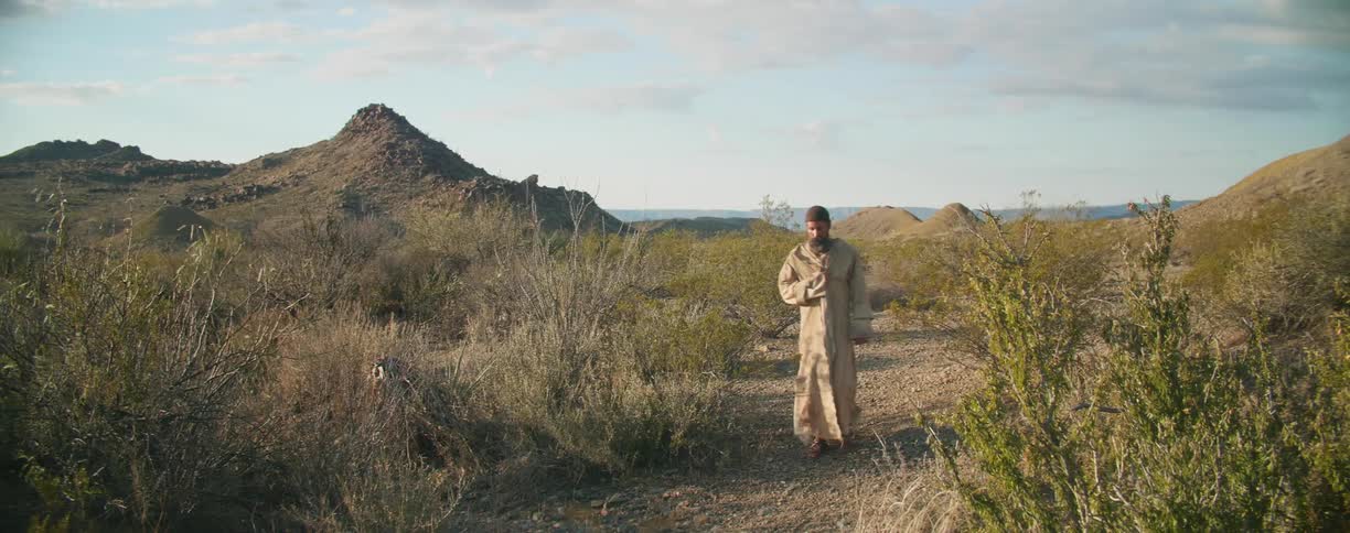 A man in robes walks through a desert on a path