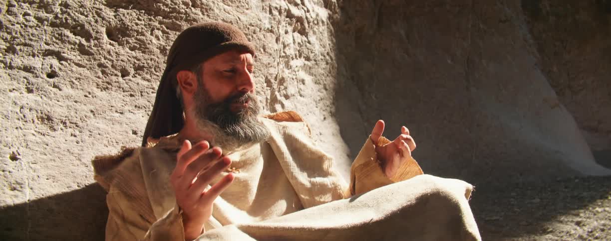 A man sitting next to a canyon wall looking at his hands then looking up to the sky