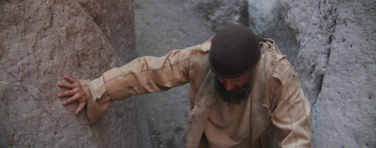 Bearded man wearing a robe climbs a narrow canyon in the desert