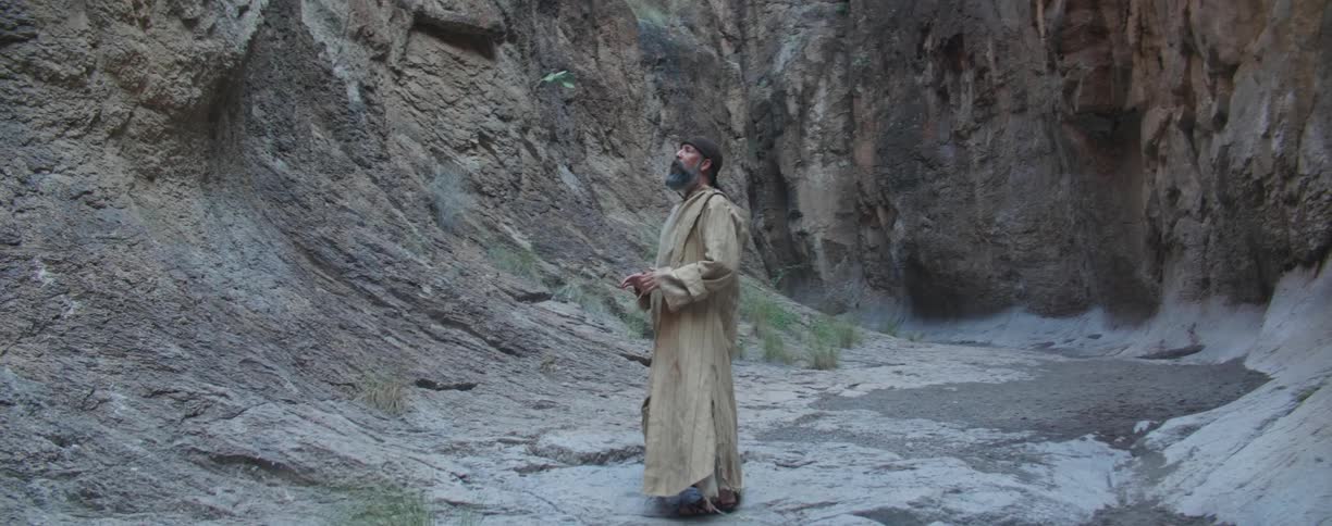 A man in a dirty robe wanders about in a narrow canyon