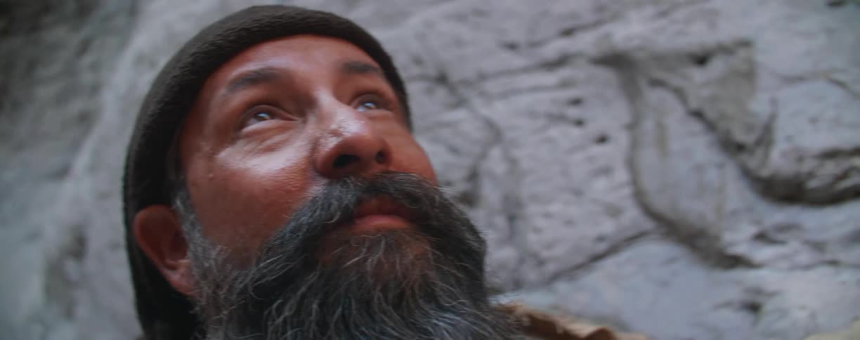 A bearded man in a headwrap gazes upwards at the sky while standing next to a large rock formation