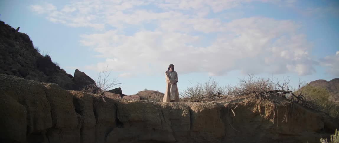 A man in a long cloak standing and looking over the edge of a cliff