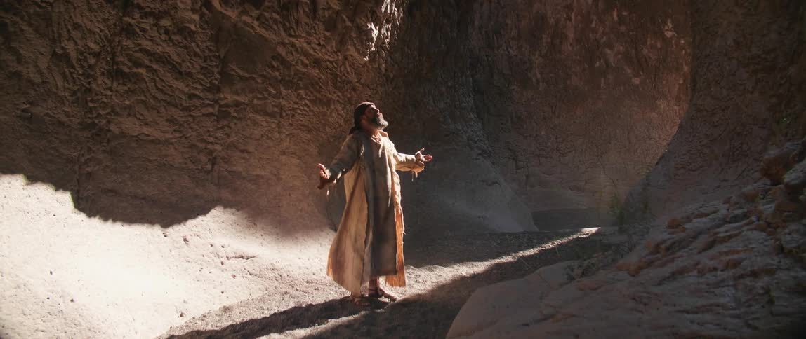 A man in a long cloak stands at the base of a canyon with his arms extended to his side
