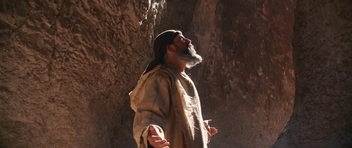 A man standing at the base of a canyon looks up to the sky with his arms extended and prays