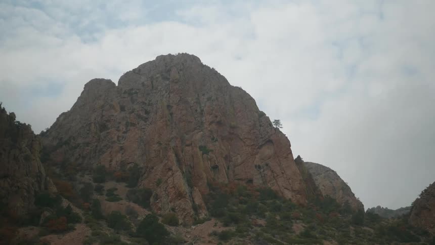 Trees stand a distance from vegetation dotted mountain composed of reddish stone