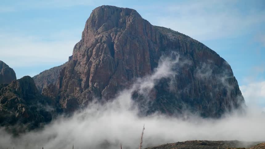 Heavy fog flows past craggy mountain peak under slightly cloudy sky