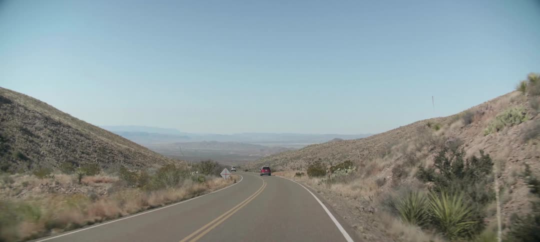 Vehicles drive down a two lane road between hills in a rural area