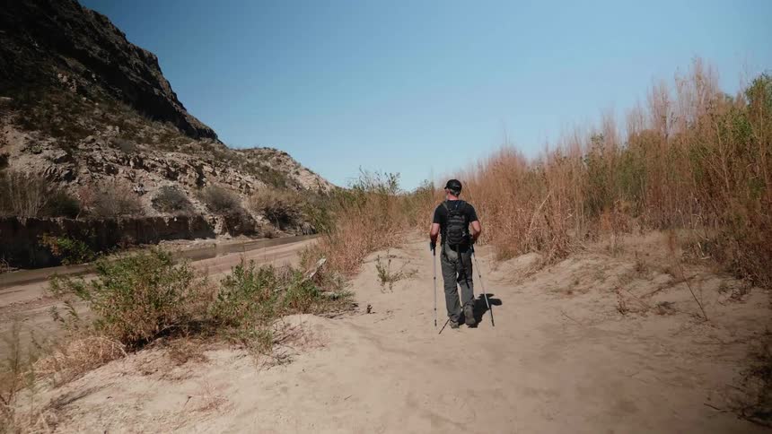 A white man carrying two metal poles strolls through a desert terrain near a mountain