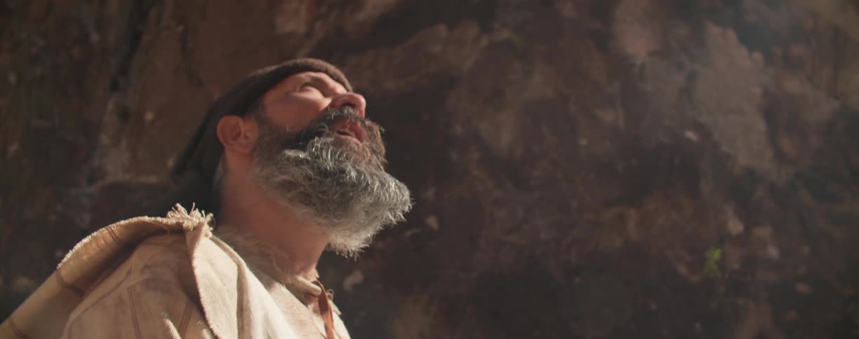 A white man with a beard and dirty clothes walks through a narrow canyon as the sun beats down on him