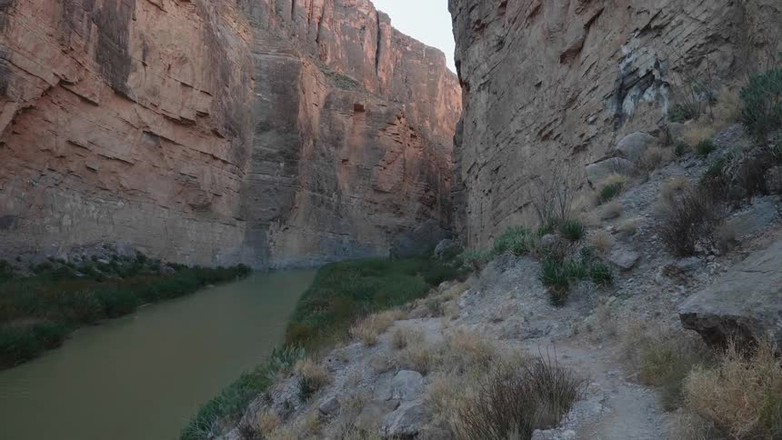 Narrow dirt path close to a river going through a canyon between tall mountain cliffs in the desert