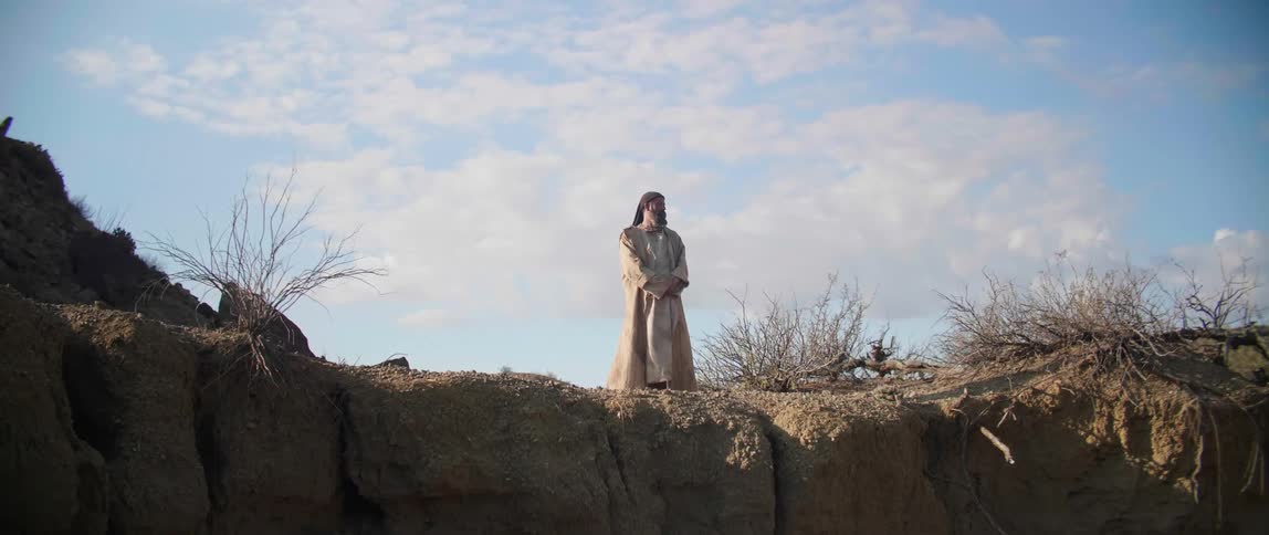 A man with a coat standing by a cliff with the sky and clouds in the background