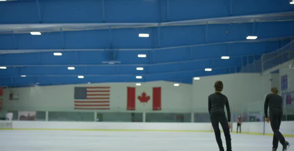 Man and woman figure skate in an arena with two flags at the end