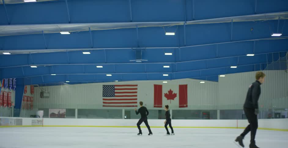 Skaters cross ice rink while others remain near wall under blue ceiling dotted with rectangular lights