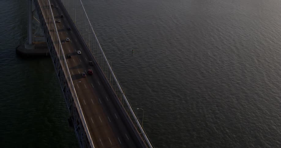 Aerial view of vehicles crossing a suspension bridge whose cables hang from towers that are attached to caissons or cofferdams with the tall buildings and skyscrapers in the background at sunset